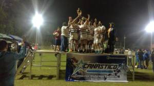 Equipo de futbol de uniforme blanco celebrando con trofeo levantado sobre tarima